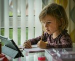 Young girl concentrating on schoolwork at home, writing in a notebook while using a tablet, illustrating learning and assessment in education.