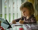 Young girl concentrating on schoolwork at home, writing in a notebook while using a tablet, illustrating learning and assessment in education.