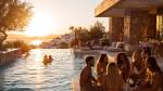 Love Island–style contestants lounge and chat beside an infinity pool at sunset outside a modern stone villa—visualizing the aspirational lifestyle that feeds the show’s popularity.