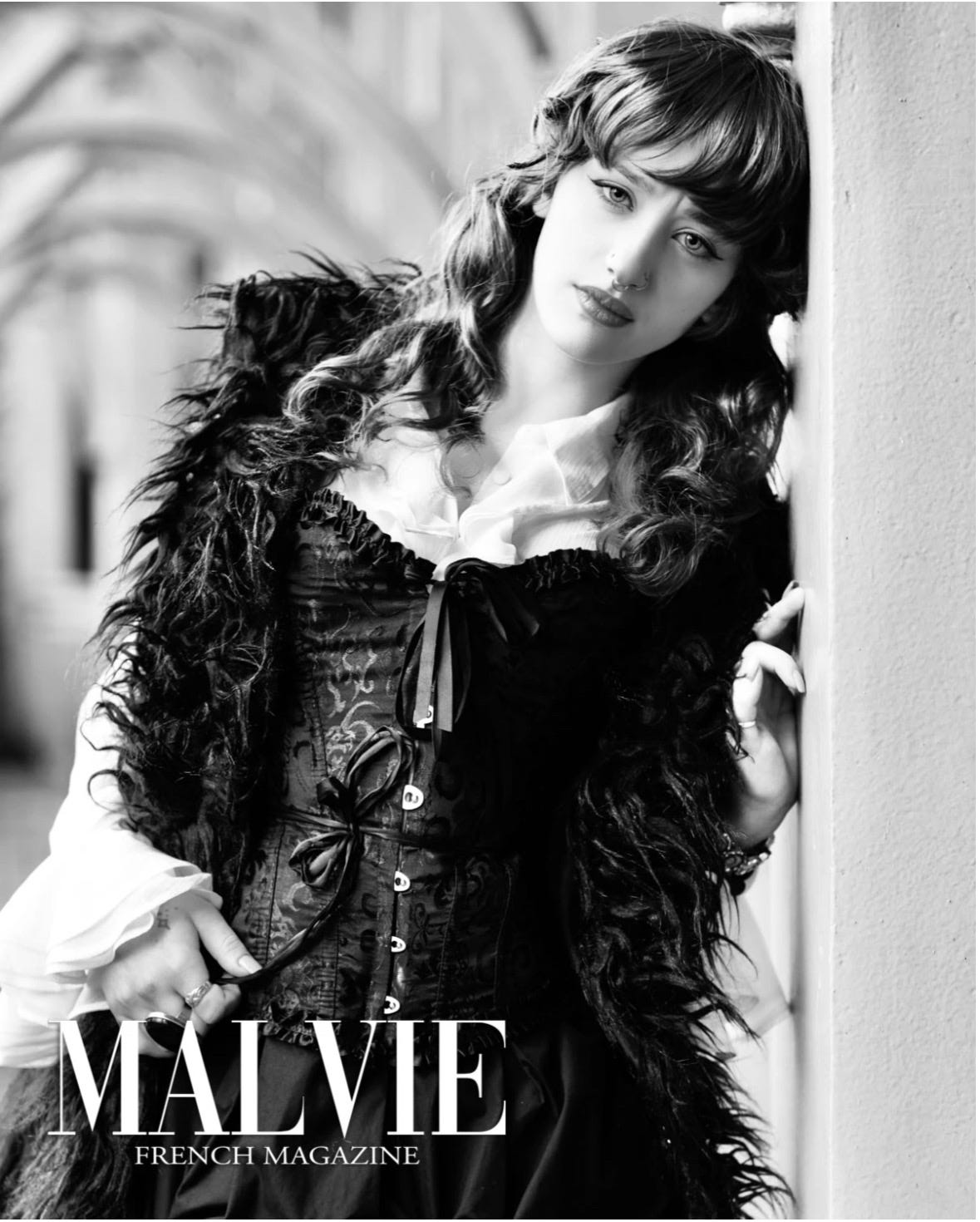 Black and white portrait of writer Abbie Veronica Davinson, featured in Malvie French Magazine, wearing a corset and textured coat, leaning against a wall with a calm, reflective expression.