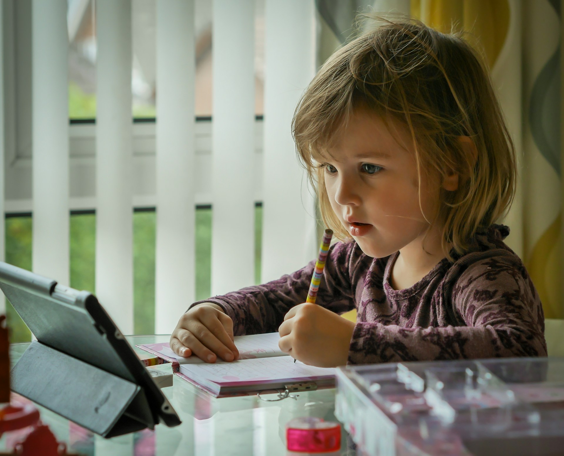 Young girl concentrating on schoolwork at home, writing in a notebook while using a tablet, illustrating learning and assessment in education.