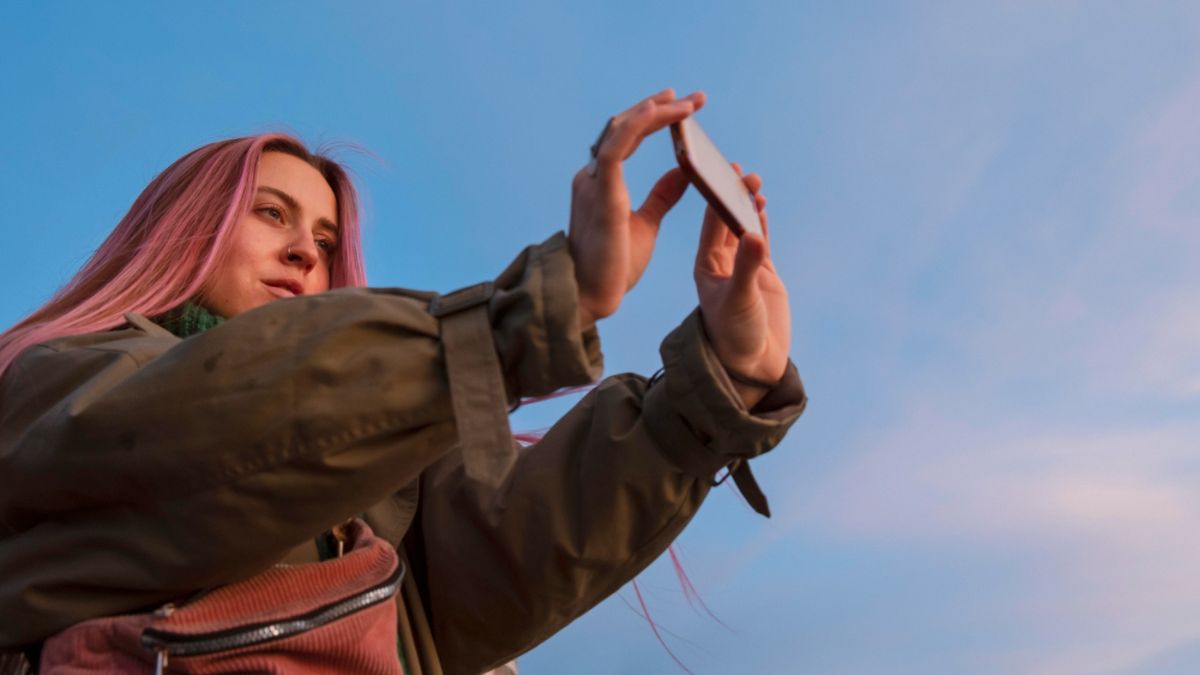 A young woman taking a photo on her phone, representing digital photo hoarding and difficulty deleting images. A young woman taking a photo on her phone, representing digital photo hoarding and difficulty deleting images.