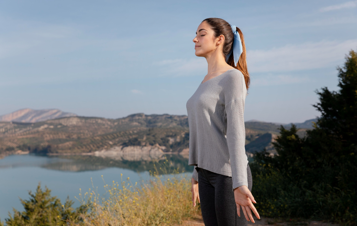 Young woman practicing mindful breathing outdoors by a lake, symbolizing the mental health benefits of exercise and emotional well-being.