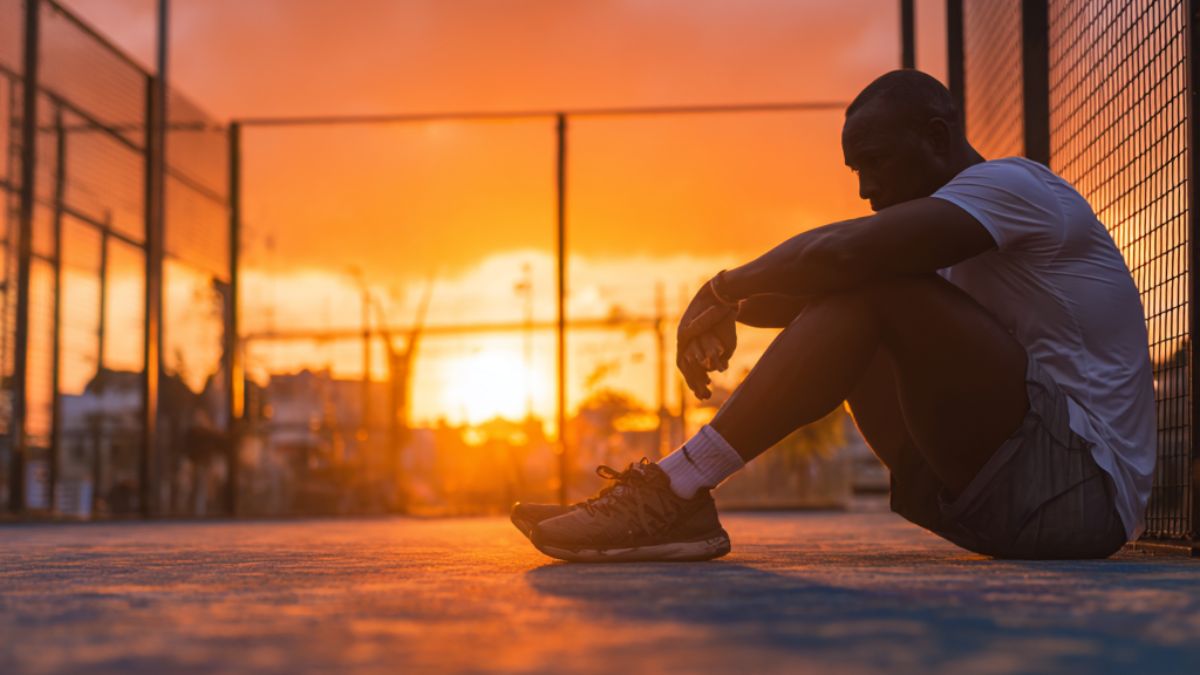 Man sitting on a padel court at sunset with his head bowed, symbolising self-reflection, frustration, and the psychology of beating yourself up after mistakes.