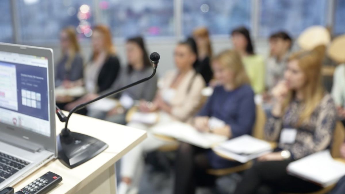 Microphone and laptop at the podium of an executive healthcare conference with attendees in the background preparing for a small-group leadership session. Microphone and laptop at the podium of an executive healthcare conference with attendees in the background preparing for a small-group leadership session.